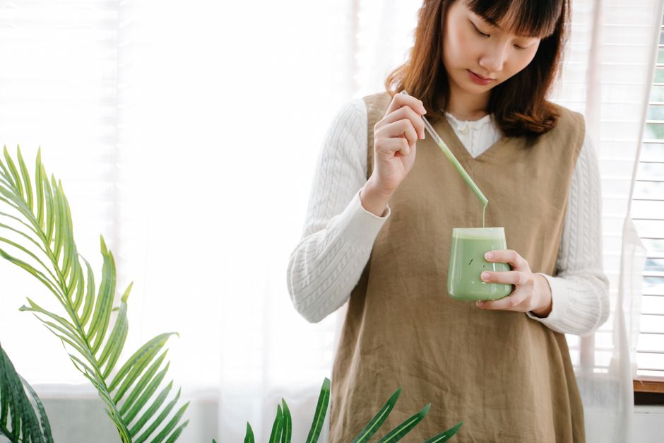 Woman stirring a vibrant green matcha beverage in a well-lit indoor setting, surrounded by lush plants.